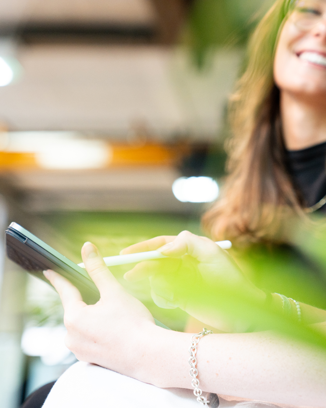 Frau mit langem braunem Haar, leicht lächelnd, bedient mit ein Tablet – im Vordergrund unscharfe grüne Pflanzen, im Hintergrund ein modernes, helles Büro.